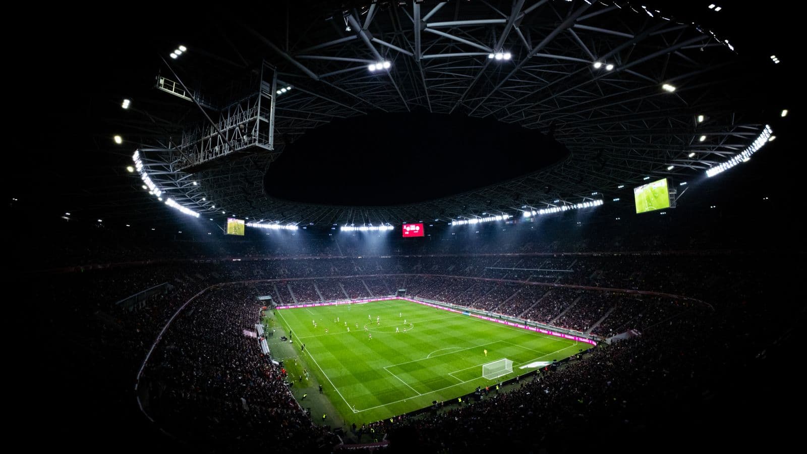 Rozświetlony stadion nocą z lotu ptaka, trybuny wypełnione kibicami