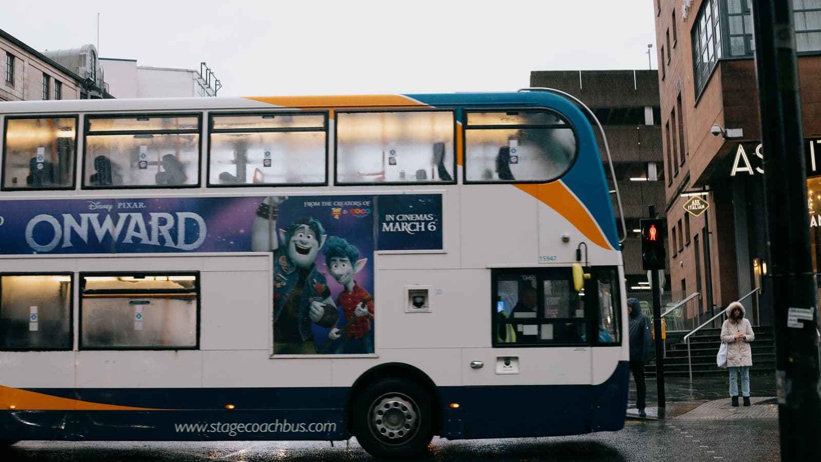 Piętrowy autobus z ilustracją jedzie mokrą ulicą obok anonimowych przechodniów